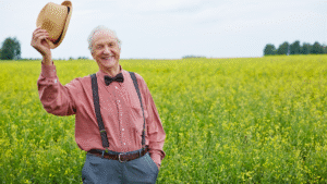 A happy senior man, wearing a red checkered shirt, bow tie, and suspenders, smiles while tipping his straw hat in a vast yellow canola field.