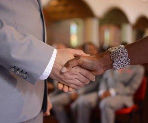 Diverse hands shake, one in a grey suit, the other adorned with a decorative silver bracelet. This signifies unity and agreement in a church setting.